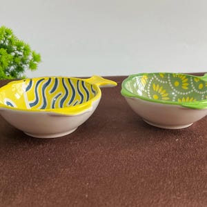 Puede incluir: Dos cuencos de cerámica con forma de pez. Uno es amarillo con líneas onduladas azules, el otro es verde con motivos florales amarillos. Ambos tienen una base blanca y una cola de pez.