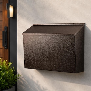 May include: A dark brown, rectangular wall-mounted mailbox with a textured, matte finish. The mailbox features a slanted top lid that extends over the front panel, creating a clean, geometric profile mounted on a light-colored exterior wall.