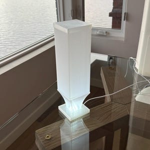 May include: A white, rectangular lamp with a ribbed design. The lamp has a square base and top, and it is illuminated from within. The lamp is sitting on a glass table with a wooden frame, near a window overlooking the water.