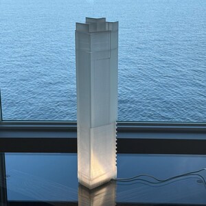 May include: A translucent, rectangular lamp resembling a skyscraper, emitting a bright internal light. The lamp is positioned on a dark surface before a window with a view of water. The lamp features a textured surface and a distinctive architectural design.
