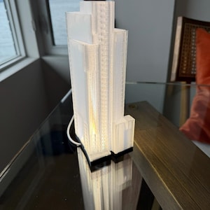 May include: A white, 3D-printed skyscraper lamp with a rectangular base and multiple tiers. The lamp is lit from within, casting a warm glow. The lamp is on a reflective surface.