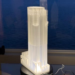 May include: A white, illuminated architectural model of a skyscraper. The model is lit from within, showcasing the building's design. It sits on a black rotating base with a power cord. The background features a body of water and a cityscape at dusk.