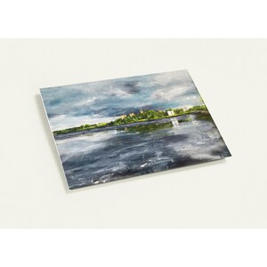 May include: A landscape print featuring a river scene under a cloudy sky. The artwork depicts a body of water reflecting the sky and a distant shoreline with buildings and trees. The color palette includes shades of blue, gray, and green.