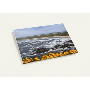 May include: A rectangular print of a coastal landscape. The image depicts a turbulent sea with dark waves crashing against black rocks. A strip of yellow-orange vegetation lines the bottom, with a sandy beach and cloudy sky in the background.