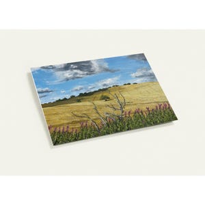 May include: A landscape painting depicts a golden field under a blue sky with fluffy clouds. A lone tree stands on a distant hill. Purple wildflowers and a bare branch are in the foreground. The artwork is on a rectangular canvas.