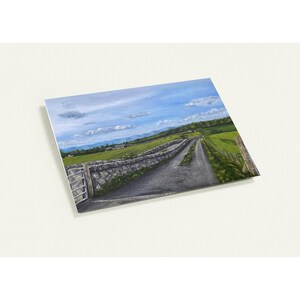 May include: An original landscape painting depicts a rural road winding through green fields under a blue sky with fluffy clouds. A stone wall and gate border the road, leading towards distant hills and buildings. The artwork is presented on a white background.
