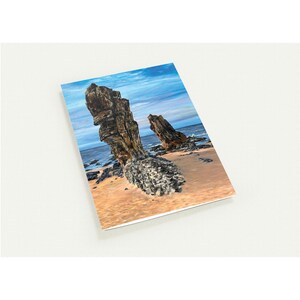May include: A notebook featuring a coastal landscape with large, weathered rock formations on a sandy beach. The sky is blue with streaks of clouds, and the ocean is visible in the background. The rocks are brown and gray.