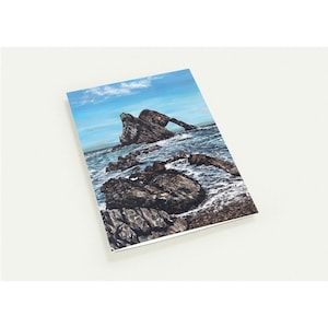 May include: A landscape art print featuring a coastal scene with a rock archway. The artwork depicts dark, textured rocks surrounded by blue water under a blue sky with clouds. The print is on a white background.