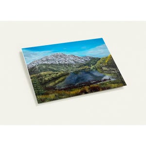 Può includere: Un dipinto di paesaggio raffigurante una catena montuosa con una vetta innevata, un lago sereno e una vegetazione verde lussureggiante. Il cielo è di un blu vibrante con nuvole sottili. L'opera d'arte è esposta su una superficie bianca.