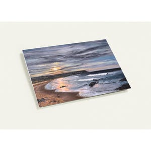 May include: An oil painting depicts a coastal scene at sunset. The artwork features a sandy beach, ocean waves, and a cloudy sky with hues of blue, gray, and orange. The painting is on a rectangular canvas.