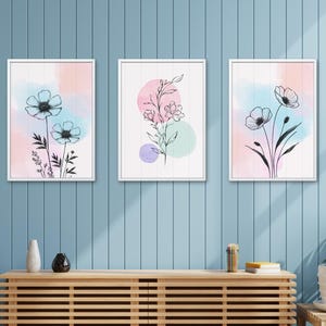 May include: Three framed floral art prints. Each print features a watercolor background in pastel colors with black line drawings of flowers and foliage. The frames are white, and the art hangs on a blue wall.