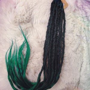 Könnte beinhalten: Ein Bündel aus dunkelgrünen und teal-farbenen Kunst-Dreadlocks. Die Dreadlocks sind oben mit einem kleinen Etikett zusammengebunden. Die Dreadlocks werden auf einer flauschigen, weißen Oberfläche präsentiert.