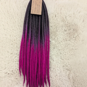 May include: A bundle of handmade dreadlocks in a gradient of colors. The top section is dark gray, transitioning to purple, and finally ending in a vibrant magenta. A tag with text is attached to the top.