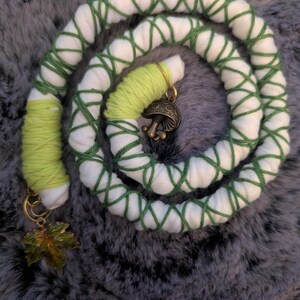 Könnte beinhalten: Ein langes, flexibles Haaraccessoire aus weißem Stoff, das mit dunkelgrünem Garn kreuz und quer umwickelt ist. Es enthält Abschnitte aus hellgrünem Garn, einen pilzförmigen Anhänger aus bronzefarbenem Metall und einen Ahornblatt-Anhänger aus Bronze.