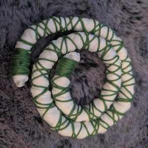 Puede incluir: Un objeto cilíndrico largo y flexible hecho de tela blanca envuelto en un patrón entrecruzado con hilo verde oscuro. Los extremos del cilindro están fuertemente enrollados con el mismo hilo verde oscuro, creando una forma de espiral.