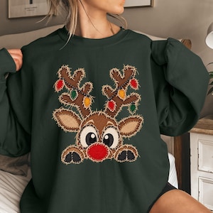 May include: A dark green crewneck sweatshirt featuring a central graphic of a brown reindeer with large eyes, a bright red nose, and antlers decorated with colorful Christmas light bulbs. The design has a textured, stitched-on appearance.