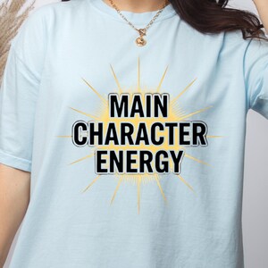 Puede incluir: Una camiseta azul claro con las palabras "MAIN CHARACTER ENERGY" en blanco y negro, sobre un diseño de rayos de sol amarillos. La camiseta está hecha de un material suave.