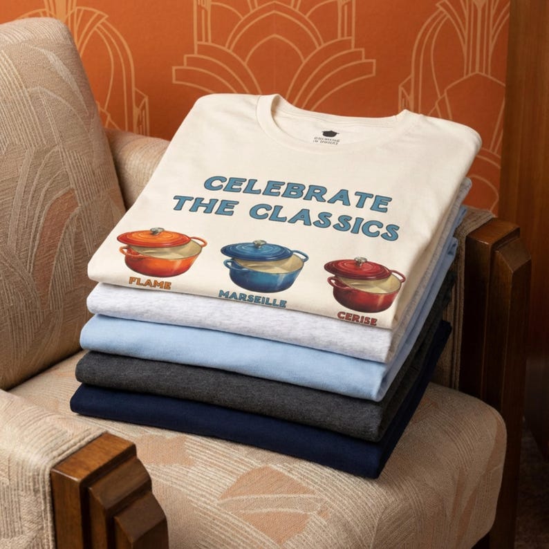A stack of folded crew-neck t-shirts sitting on a patterned upholstered chair. Top t-shirt reads, "CELEBRATE THE CLASSICS" with three dutch ovens, in colors Flame, Marseille, and Cerise.