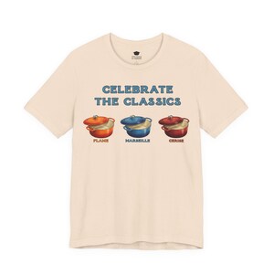 Cream colored crew-neck t-shirt reads, "CELEBRATE THE CLASSICS" with three dutch ovens, in colors Flame, Marseille, and Cerise.