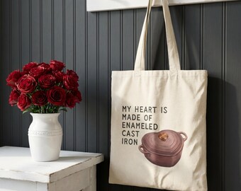 Puede incluir: Un bolso tote color crema con el texto "MY HEART IS MADE OF ENAMELED CAST IRON" y un gráfico de una olla de hierro fundido marrón en forma de corazón. La bolsa cuelga de una pared blanca. Un ramo de rosas rojas en un jarrón blanco.