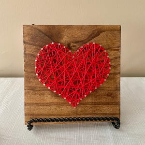 May include: A wooden heart string art piece. The heart is made of red string and is mounted on a stained wooden block. The piece is supported by a black metal stand. The background is a neutral color.