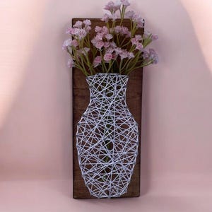 以下が含まれることがあります： 白い糸アートで作られた花瓶の形をした木製の壁掛け。花瓶には、緑の茎を持つピンクの花のブーケが入っています。木製のベースは長方形で、濃い茶色に着色されています。