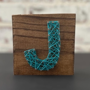 May include: Wooden block with a teal string art letter "J". The letter is crafted with teal thread wrapped around small silver nails, creating a textured design. The wood has a dark brown stain.