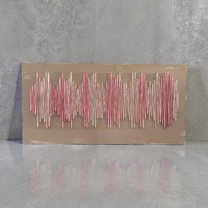May include: A rectangular wooden wall art piece with a beige background. Pink and white thread is strung across the board in a wave-like pattern, creating a textured design. The artwork is displayed against a gray wall.