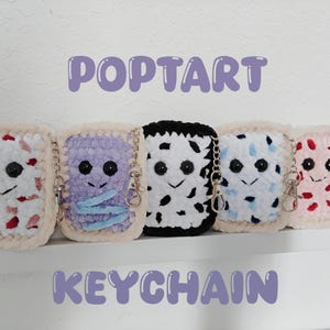 May include: Five handmade crochet Pop-Tart keychains in various colors, including white with red sprinkles, purple, black and white, blue and white, and pink with red sprinkles. Each keychain has a smiling face and a silver clasp.