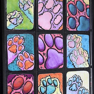 May include: Nine colorful paw print illustrations in a black card holder. Each card features a unique paw print design with vibrant colors like pink, orange, blue, and purple. The artwork is hand-drawn with black outlines.