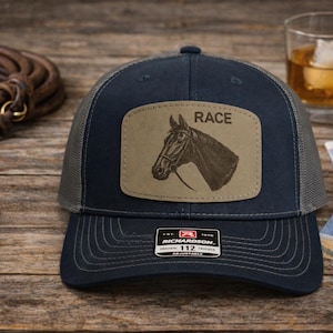 May include: A navy blue and grey trucker hat featuring a rectangular tan leather patch on the front. The patch displays an engraved horse head profile and the word RACE. A black sticker on the brim reads RICHARDSON 112 ADJUSTABLE.