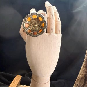May include: A vintage ring with a floral design. The ring has a silver-coloured metal setting with an amber-coloured centre and accents. It is displayed on a wooden hand mannequin against a dark background.