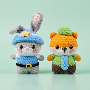 May include: Two handmade crochet stuffed animals: a gray bunny wearing a blue hat with a yellow star and a blue shirt, and an orange bear wearing a green hat, green shirt, and blue tie. The toys have black eyes and pink cheeks.