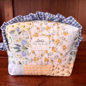 Könnte beinhalten: Eine Kosmetiktasche aus Patchwork mit blauem Vichy-Rüschenbesatz. Die Tasche hat Blumen- und geometrische Muster in Gelb, Blau und Weiß. Ein kleines Etikett lautet "Three Tiny Threads Co."