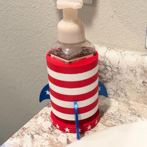 May include: A clear soap dispenser with a white pump is encased in a red, white, and blue rocket-shaped holder. The holder features red and white stripes, blue fins, and white stars, creating a patriotic theme.