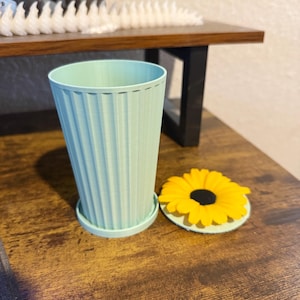 May include: A light green, ribbed planter with a matching saucer. A yellow and black sunflower coaster sits next to the planter. The items are on a wooden surface. The planter is approximately 5 inches tall.