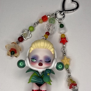 May include: A doll keychain featuring a blonde-haired figure in a green and red outfit, with a heart-shaped clasp. The keychain has beaded accents in various colors, including red, green, and yellow, and a flower charm.