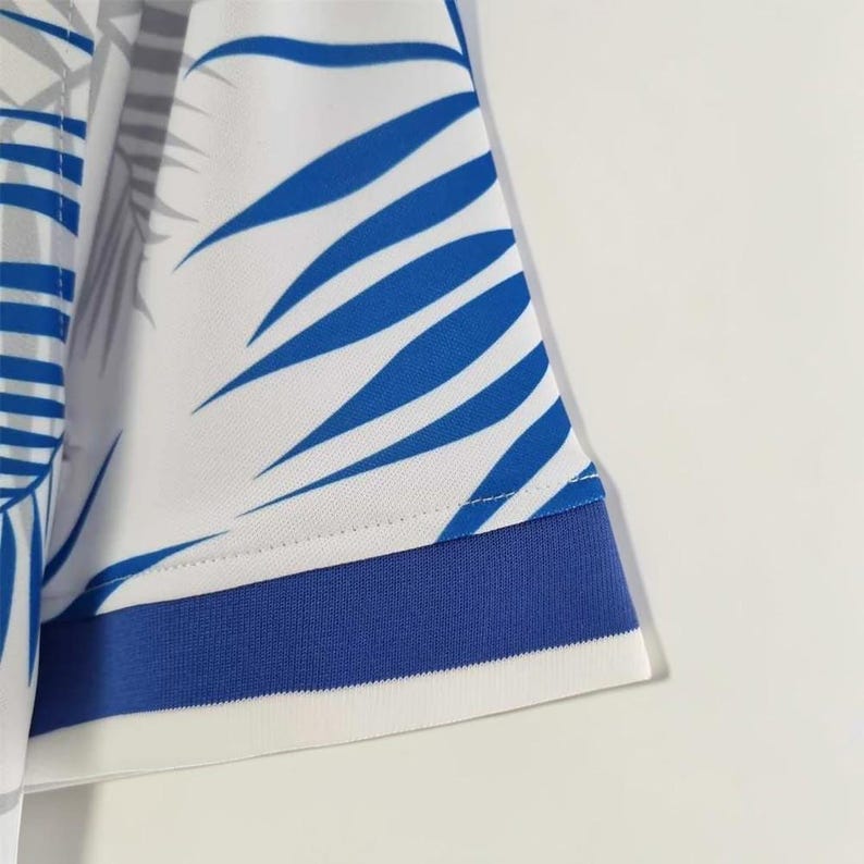 May include: Close-up of a white and blue patterned shirt sleeve. The sleeve features a blue stripe at the cuff and a design of blue leaf-like shapes on a white background. The fabric appears to be a lightweight material.