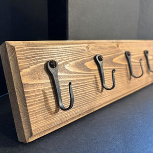May include: A rustic wooden coat rack with five black metal hooks. The wood has a warm, natural finish, and the hooks are evenly spaced along the length of the board. The hooks are attached with screws. This item is suitable for entryways or bedrooms.