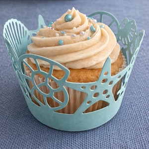 Puede incluir: Un cupcake con glaseado color crema y chispas azules y perladas se encuentra en un envoltorio para cupcakes azul claro con temática de conchas marinas. El envoltorio presenta diseños recortados de estrellas de mar y conchas. El cupcake está sobre una superficie azul.