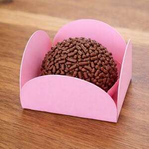 May include: A chocolate truffle covered in chocolate sprinkles sits in a pink paper candy cup. The truffle is round and dark brown, contrasting with the light pink of the cup. The candy cup is on a wooden surface.