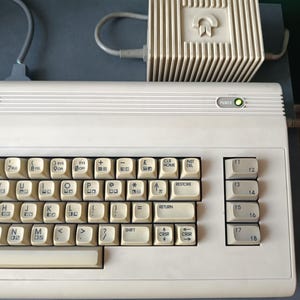 Può includere: Una tastiera per computer vintage bianco sporco con un alimentatore abbinato. La tastiera ha numerosi tasti con simboli e testo, tra cui una spia "POWER". L'alimentatore presenta un logo e un design a coste.