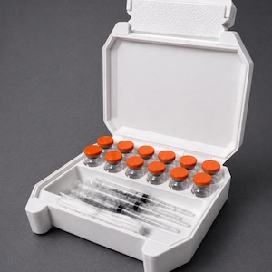 May include: A white plastic medical case, open to reveal vials with orange caps and several syringes. The case has a hinged lid and a tray to hold the contents. The background is a solid gray.