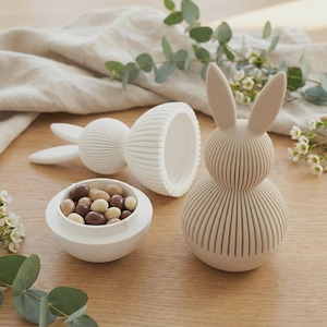 May include: A white, ridged bunny-shaped container with a matching bowl filled with chocolate-covered candies. The bunny is in three parts: the head with ears, the body, and a separate lid. The items are on a wooden surface.