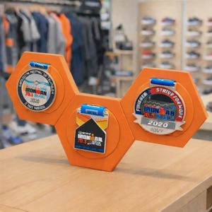 Puede incluir: Un expositor de medallas hexagonal naranja con tres medallas de finalista Ironman 70.3 Brasil 2020. Las medallas son plateadas y azules, con el texto "FINISHER STRIVE FOR MORE" y "IRONMAN 70.3 BRASIL 2020". El expositor está sobre una superficie de madera.
