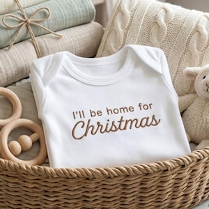 Puede incluir: Un body blanco para bebé con el texto bordado "I'll be home for Christmas" en marrón. El body está en una cesta de mimbre con un juguete de madera, un cordero de peluche y mantas dobladas.