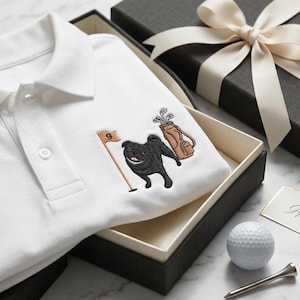Peut inclure: Un polo blanc avec une broderie représentant un carlin noir debout à côté d'un drapeau de golf marqué du chiffre 9 et d'un sac de golf marron rempli de clubs. Le polo est présenté dans une boîte cadeau noire avec un ruban crème.