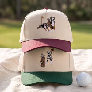 Könnte beinhalten: Zwei übereinander gestapelte Baseballkappen mit cremefarbenen Kronen. Die obere Kappe hat einen weinroten Schirm und eine Stickerei von zwei Hunden mit Golftasche und Fahne. Die untere Kappe hat einen dunkelgrünen Schirm und eine Stickerei eines Dalmatiners.