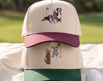 May include: Two stacked baseball caps with cream-colored crowns. The top cap has a maroon brim and an embroidered design of two dogs with a golf bag and flag. The bottom cap has a dark green brim and an embroidered design of a dalmatian with a golf bag and flag.