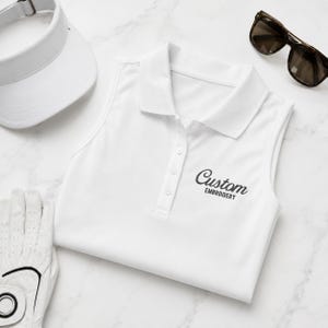 May include: A white sleeveless polo shirt with a collar and a button placket, featuring the black text Custom EMBROIDERY on the chest. The shirt is accompanied by a white visor, a white golf glove, and dark-tinted sunglasses with black frames.